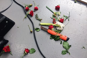 An assortment of red roses and white flowers are scattered across a gray floor. There are stems, leaves, and small pieces of greenery dispersed around. A large knife with an orange handle lies among the flowers. A black cable snakes across the floor from the left side.