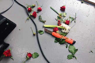 An assortment of red roses and white flowers are scattered across a gray floor. There are stems, leaves, and small pieces of greenery dispersed around. A large knife with an orange handle lies among the flowers. A black cable snakes across the floor from the left side.