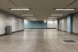 A large, empty indoor space with a tiled floor and fluorescent ceiling lights. The walls are mostly light gray and blue, with a trash can on the left side. There are several emergency exit signs on the walls and a large window at the far end of the room.