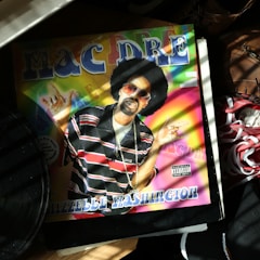 An album cover featuring a person with sunglasses and an afro hairstyle, wearing a black and red striped shirt. The background is a colorful rainbow pattern with bold lettering at the top. Surrounding objects include a vinyl record and a sneaker.