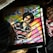 An album cover featuring a person with sunglasses and an afro hairstyle, wearing a black and red striped shirt. The background is a colorful rainbow pattern with bold lettering at the top. Surrounding objects include a vinyl record and a sneaker.