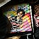 An album cover featuring a person with sunglasses and an afro hairstyle, wearing a black and red striped shirt. The background is a colorful rainbow pattern with bold lettering at the top. Surrounding objects include a vinyl record and a sneaker.
