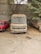 An old, dusty van is parked in an outdoor area between a dilapidated red car and a beige vehicle. The vehicles are situated under a sparse tree and against a concrete wall, with leaves and debris scattered around the ground.