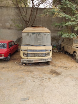 An old, dusty van is parked in an outdoor area between a dilapidated red car and a beige vehicle. The vehicles are situated under a sparse tree and against a concrete wall, with leaves and debris scattered around the ground.