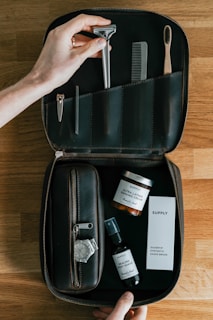 A neatly organized leather toiletry bag contains grooming items placed on a wooden surface. It holds a razor, comb, toothbrush, nail clipper, tweezers, a jar of shaving cream, a spray bottle labeled 'Healing Post Shave,' and a box labeled 'Silvertip Synthetic Shave Brush.' A wristwatch is placed next to the bag.