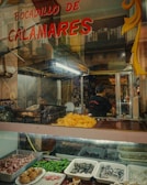 A food stall with various seafood and ingredients displayed, including sliced squid, peppers, and shellfish. The glass counter has trays filled with different types of food items. Above, a sign reads 'Bocadillo de Calamares'. Inside, people are preparing food in a bustling atmosphere.