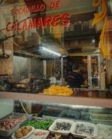 A food stall with various seafood and ingredients displayed, including sliced squid, peppers, and shellfish. The glass counter has trays filled with different types of food items. Above, a sign reads 'Bocadillo de Calamares'. Inside, people are preparing food in a bustling atmosphere.