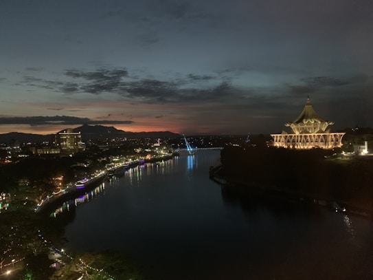 A calm river at dusk with a cityscape featuring a large, illuminated building with unique architecture on the right side. The sky transitions from warm tones near the horizon to darker shades as it rises. Surrounding buildings and bridges are adorned with lights that reflect in the water.