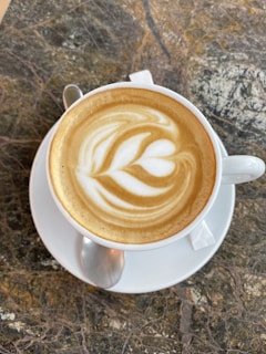 A steaming cup of cappuccino with delicate latte art resting on a saucer beside a small spoon.