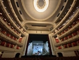An ornate and expansive theater interior with luxurious red seats and balconies, highlighted by an elaborate ceiling design featuring a grand chandelier. The stage is set with a backdrop resembling a crumbling building facade.