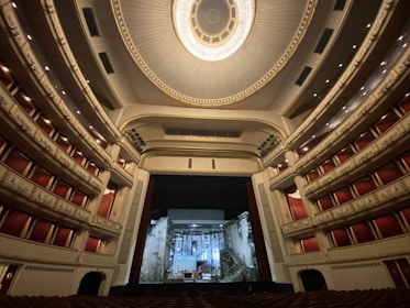 An ornate and expansive theater interior with luxurious red seats and balconies, highlighted by an elaborate ceiling design featuring a grand chandelier. The stage is set with a backdrop resembling a crumbling building facade.