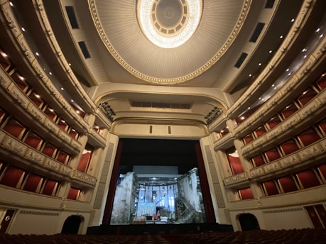 An ornate and expansive theater interior with luxurious red seats and balconies, highlighted by an elaborate ceiling design featuring a grand chandelier. The stage is set with a backdrop resembling a crumbling building facade.