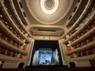 An ornate and expansive theater interior with luxurious red seats and balconies, highlighted by an elaborate ceiling design featuring a grand chandelier. The stage is set with a backdrop resembling a crumbling building facade.