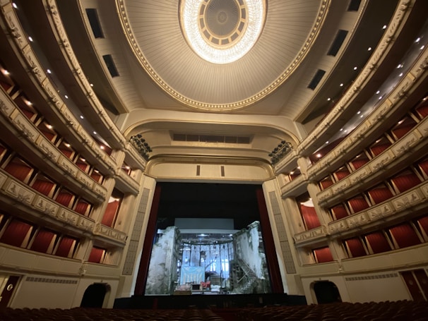 An ornate and expansive theater interior with luxurious red seats and balconies, highlighted by an elaborate ceiling design featuring a grand chandelier. The stage is set with a backdrop resembling a crumbling building facade.
