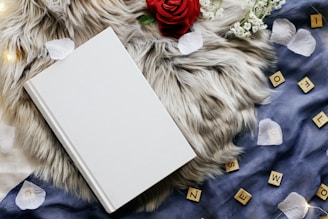 A blank white hardcover book lies on a soft, fluffy gray surface. Surrounding it are a red rose, scattered white flower petals, small white flowers, and wooden Scrabble tiles on a dark blue fabric.