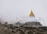 A stupa with a golden spire is situated on a rugged hillside, surrounded by mist and adorned with colorful prayer flags. Three figures holding walking sticks approach from the left, dressed in hiking gear, indicating a mountainous trek.