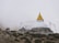 A stupa with a golden spire is situated on a rugged hillside, surrounded by mist and adorned with colorful prayer flags. Three figures holding walking sticks approach from the left, dressed in hiking gear, indicating a mountainous trek.