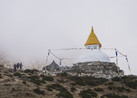 A stupa with a golden spire is situated on a rugged hillside, surrounded by mist and adorned with colorful prayer flags. Three figures holding walking sticks approach from the left, dressed in hiking gear, indicating a mountainous trek.