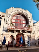 The facade of an old building with ornate architecture features a large circular window and a red wrought iron balcony railing. The name 'Abel Pereira da Fonseca' is prominently displayed above the window. Blue umbrellas and outdoor tables are set up on the cobblestone street below, with a person walking past.