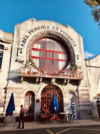 The facade of an old building with ornate architecture features a large circular window and a red wrought iron balcony railing. The name 'Abel Pereira da Fonseca' is prominently displayed above the window. Blue umbrellas and outdoor tables are set up on the cobblestone street below, with a person walking past.