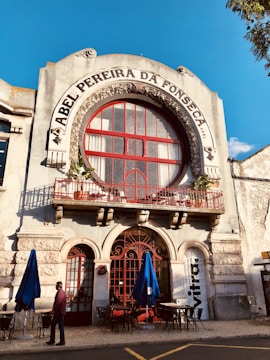 The facade of an old building with ornate architecture features a large circular window and a red wrought iron balcony railing. The name 'Abel Pereira da Fonseca' is prominently displayed above the window. Blue umbrellas and outdoor tables are set up on the cobblestone street below, with a person walking past.