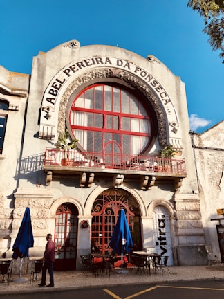 The facade of an old building with ornate architecture features a large circular window and a red wrought iron balcony railing. The name 'Abel Pereira da Fonseca' is prominently displayed above the window. Blue umbrellas and outdoor tables are set up on the cobblestone street below, with a person walking past.