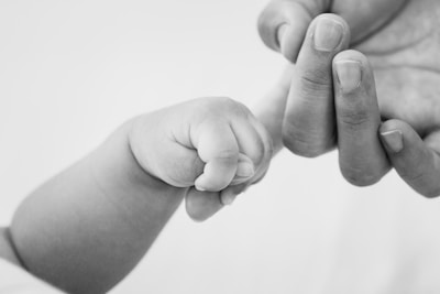 A close-up image captures an infant's small hand gently grasping an adult's finger. The scene is intimate and tender, highlighting the contrast in size between the two hands.