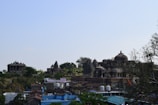 Ancient stone temples and structures sit amidst lush greenery, with a small village in the foreground featuring rustic rooftops and vibrant blue and red accents. The architecture includes ornate carvings and dome-like features typical of historical religious sites.