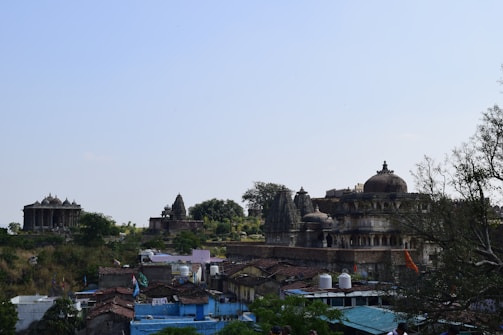Ancient stone temples and structures sit amidst lush greenery, with a small village in the foreground featuring rustic rooftops and vibrant blue and red accents. The architecture includes ornate carvings and dome-like features typical of historical religious sites.