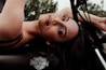A woman is reclining on a Harley-Davidson motorcycle, with her head tilted back and eyes looking towards the camera. Her dark hair cascades over the motorcycle, and she wears makeup that emphasizes her eyes and lips. The background features some blurred greenery.