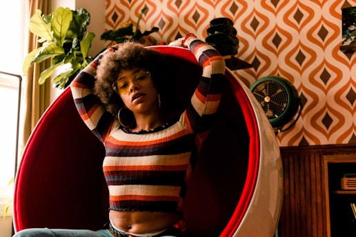 A person with an afro hairstyle is sitting in a modern, red, egg-shaped chair. They are wearing a striped long-sleeve shirt with various earthy tones. The background features an orange and white retro patterned wallpaper, a large green plant, and a vintage fan. The space has a warm, vintage aesthetic.