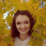 A woman wrapped in a bright yellow scarf smiling against a backdrop of autumn leaves.
