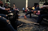 People seated in an airport lounge, some using electronic devices, with luggage around. A small dog is also present on the floor wearing a pink harness. The flooring has a patterned design, and the lighting is dim.