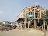 A multi-story building under construction with wooden scaffolding and several workers spread across different levels. The environment appears busy with construction materials, including cement bags and tools on the ground. A blue sky is visible above, and a few residential houses can be seen in the background.