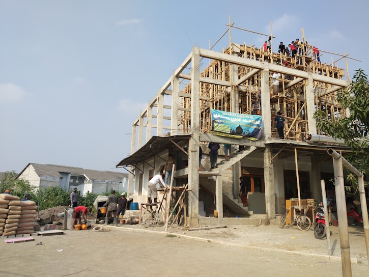 A multi-story building under construction with wooden scaffolding and several workers spread across different levels. The environment appears busy with construction materials, including cement bags and tools on the ground. A blue sky is visible above, and a few residential houses can be seen in the background.