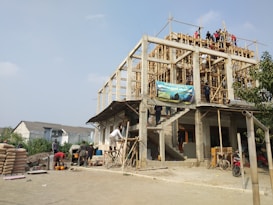 A multi-story building under construction with wooden scaffolding and several workers spread across different levels. The environment appears busy with construction materials, including cement bags and tools on the ground. A blue sky is visible above, and a few residential houses can be seen in the background.