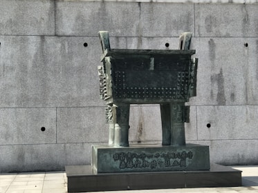 A large bronze sculpture resembling an ancient Chinese ritual vessel stands against a plain concrete wall. The vessel features intricate carvings and two prominent handles on either side. The surface appears weathered, adding an aged and historical feel to the artifact.