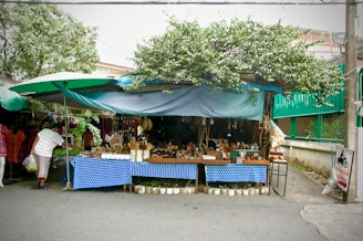 A small outdoor market stall under a leafy tree, with a blue polka-dotted tablecloth covering the display tables. The tables are filled with various handcrafted items and wooden utensils. A person is browsing the items on the left side of the stall. The stall is sheltered with tarps, and there are other shops with clothes visible in the background. The street is quiet, and there is a sign with Thai writing on the side.