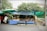 A small outdoor market stall under a leafy tree, with a blue polka-dotted tablecloth covering the display tables. The tables are filled with various handcrafted items and wooden utensils. A person is browsing the items on the left side of the stall. The stall is sheltered with tarps, and there are other shops with clothes visible in the background. The street is quiet, and there is a sign with Thai writing on the side.