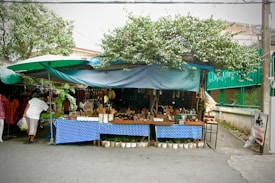 A small outdoor market stall under a leafy tree, with a blue polka-dotted tablecloth covering the display tables. The tables are filled with various handcrafted items and wooden utensils. A person is browsing the items on the left side of the stall. The stall is sheltered with tarps, and there are other shops with clothes visible in the background. The street is quiet, and there is a sign with Thai writing on the side.
