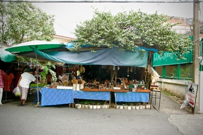 A small outdoor market stall under a leafy tree, with a blue polka-dotted tablecloth covering the display tables. The tables are filled with various handcrafted items and wooden utensils. A person is browsing the items on the left side of the stall. The stall is sheltered with tarps, and there are other shops with clothes visible in the background. The street is quiet, and there is a sign with Thai writing on the side.