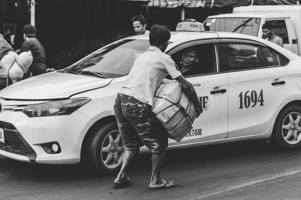 fotografia in scala di grigi di un uomo che trasporta una scatola di cartone accanto al taxi