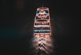 A well-lit boat cruises through dark waters, leaving a glowing trail. The vessel is adorned with strings of lights, creating an inviting and festive atmosphere. The top deck features a canopy and there appear to be people on board enjoying the ambiance.