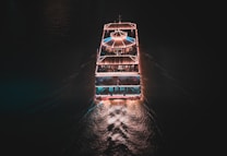 A well-lit boat cruises through dark waters, leaving a glowing trail. The vessel is adorned with strings of lights, creating an inviting and festive atmosphere. The top deck features a canopy and there appear to be people on board enjoying the ambiance.