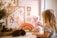 A child interacts with a pink teddy bear on a table adorned with a wooden rainbow and cloud decoration. In the background, there are framed pieces of floral artwork with earthy colors. Natural elements like pine cones and dried flowers in a vase add an organic touch to the scene.