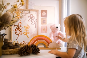 A child interacts with a pink teddy bear on a table adorned with a wooden rainbow and cloud decoration. In the background, there are framed pieces of floral artwork with earthy colors. Natural elements like pine cones and dried flowers in a vase add an organic touch to the scene.