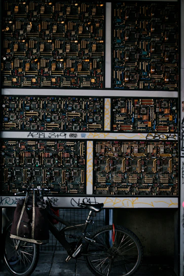 A wall decorated with multiple circuit boards, showcasing intricate electronic components, is adorned with graffiti. In front of the wall, a bicycle with a bag attached to it is parked, creating a blend of urban and technological elements.