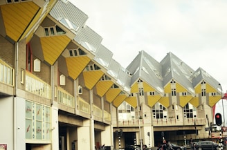 A series of uniquely designed cube-shaped buildings with a vibrant yellow and gray color scheme. The structures are elevated, forming an architectural complex that appears to defy conventional gravity with their tilted orientation. The scene includes a few pedestrians and vehicles passing by in an urban setting.
