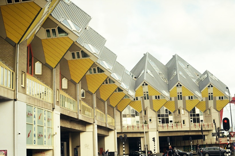 A series of uniquely designed cube-shaped buildings with a vibrant yellow and gray color scheme. The structures are elevated, forming an architectural complex that appears to defy conventional gravity with their tilted orientation. The scene includes a few pedestrians and vehicles passing by in an urban setting.