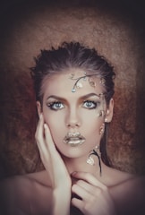 A striking portrait of a person with intricate gold and silver metallic makeup. The adornments resemble delicate leaves and branches, highlighting the eyes and lips. The background is a soft brown, enhancing the overall dramatic effect with a mix of natural and surreal elements.